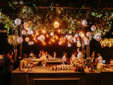 Weselny drink bar pod zielenią i kulistymi lampami, barmani przygotowują drinki dla gości