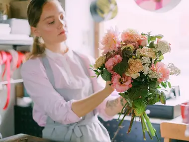 Florystka w fartuchu układa w pracowni bukiet z różowych dalii, goździków i zieleni