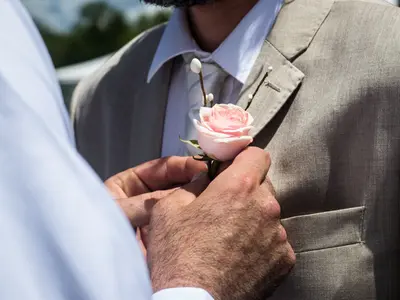 Dłonie przypinają butonierkę z różowej róży do klapy beżowej marynarki Pana Młodego