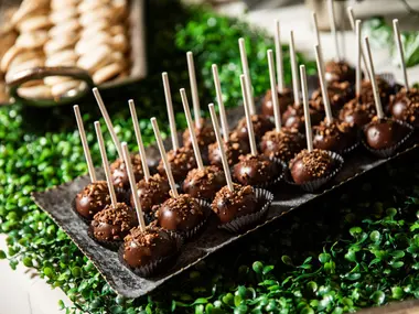 Czekoladowe cake pops na patyczkach ułożone na tacy wśród zieleni na słodkim stole