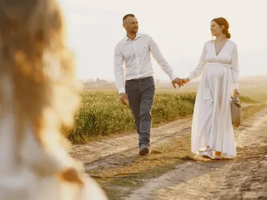 Para w ciąży spaceruje za rękę polną drogą, kobieta w długiej białej sukni