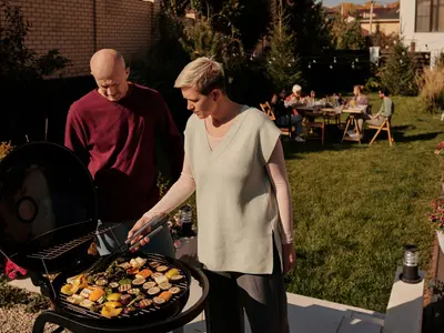 Para griluje warzywa na okrągłym grillu w ogrodzie, w tle goście przy stołach na trawie