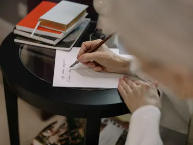 Dłoń pisze piórem dedykację na kartce przy stoliku, obok leżą książki
