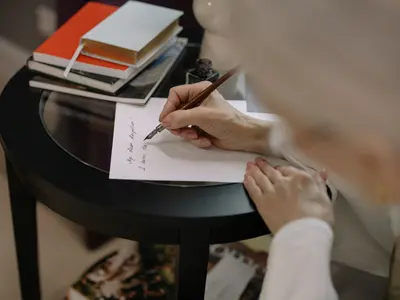 Dłoń pisze piórem dedykację na kartce przy stoliku, obok leżą książki