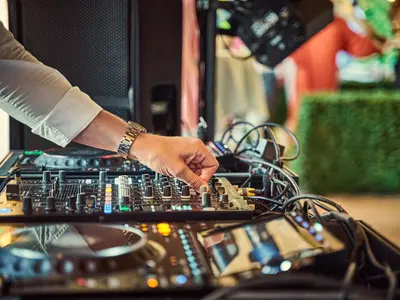 Dłoń DJ-a reguluje mikser na konsoli podczas przyjęcia, w tle zieleń i goście