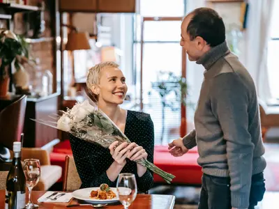 Uśmiechnięta kobieta przy stole w restauracji dostaje od męża bukiet białych róż