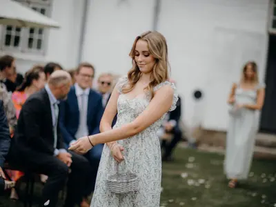 Młoda kobieta w kwiecistej sukience sypie płatki przed gośćmi ceremonii w plenerze