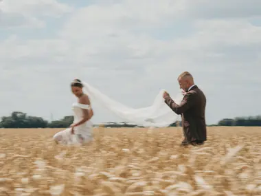 Pan młody unosi długi welon panny młodej idącej przez złote pole zboża
