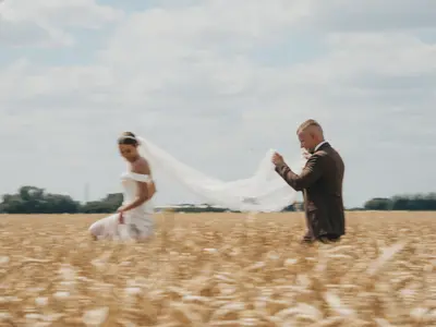 Pan młody unosi długi welon panny młodej idącej przez złote pole zboża