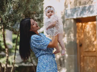 Mama w niebieskiej sukience unosi niemowlę w koronkowej sukience z opaską