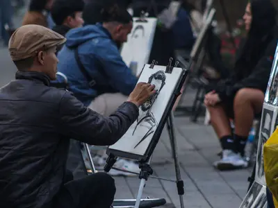 Uliczny rysownik w kaszkiecie szkicuje portret na sztaludze wśród innych artystów