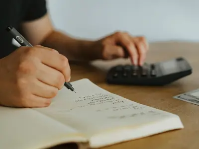 Dłonie zapisują wydatki w notesie i liczą na kalkulatorze przy drewnianym stole