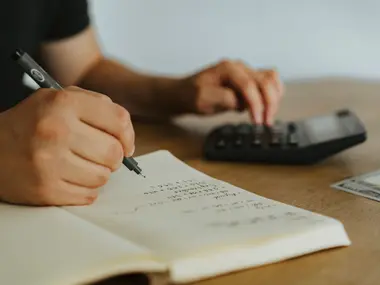 Dłonie zapisują wydatki w notesie i liczą na kalkulatorze przy drewnianym stole