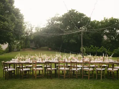 Długi stół weselny nakryty w ogrodzie, ze świecami, kwiatami i girlandami lampek nad trawnikiem