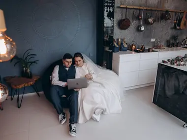 Para młoda w stroju ślubnym siedzi razem w nowoczesnej kuchni i ogląda coś na laptopie