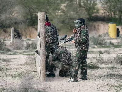 Gracze w maskach i moro kryją się za drewnianym słupem na polu do paintballa