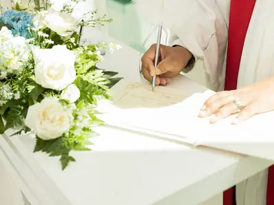Dłoń z obrączką wpisuje się długopisem do białej księgi obok bukietu białych róż