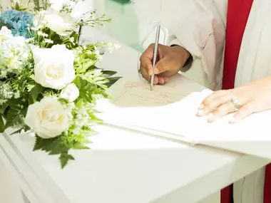 Dłoń z obrączką wpisuje się długopisem do białej księgi obok bukietu białych róż