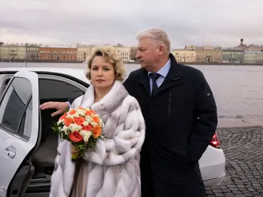 Dojrzała Panna Młoda w futrze z bukietem pomarańczowych róż stoi z mężem przy samochodzie