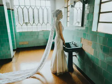 Panna Młoda w koronkowej sukni syrence z długim welonem stoi w łazience z zielonymi kafelkami