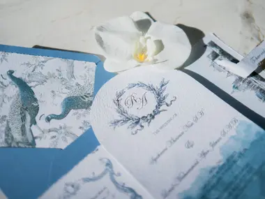Niebieskie zaproszenia ślubne z motywem pawi i monogramem obok białej orchidei