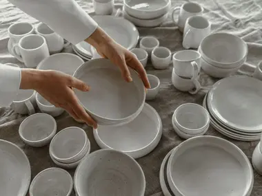 Dłonie układają białą ceramiczną zastawę: talerze, miski i kubki na lnianym obrusie