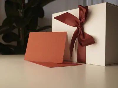 Kremowe pudełko z bordową wstążką i czerwoną kartką z kopertą na stole