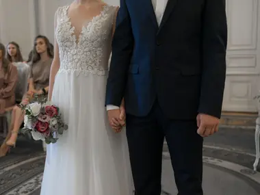 Para młoda trzyma się za ręce w eleganckiej sali, w tle siedzą goście