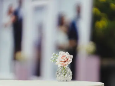 Pojedyncza pudroworóżowa róża w kryształowym wazoniku na białym stole, w tle rozmyte sylwetki
