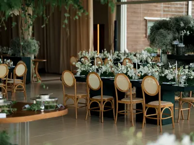 Sala weselna z ciemnozielonymi stołami, rattanowymi krzesłami i białymi kwiatami wśród zieleni