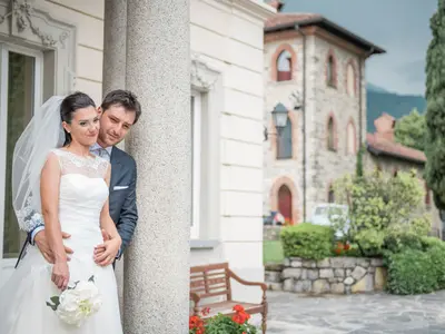 Pan młody obejmuje pannę młodą przy kolumnie jasnej willi, w tle kamienny budynek