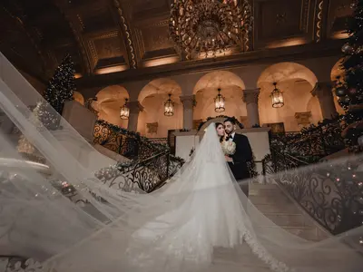 Para młoda na schodach eleganckiej sali z choinkami, długi welon rozpostarty na pierwszym planie