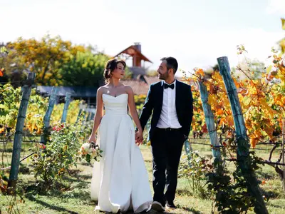 Para młoda idzie za rękę przez winnicę, wśród liści winorośli zaczynających się złocić