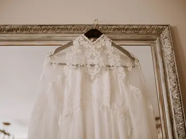 Koronkowa suknia ślubna z wysokim kołnierzem wisi na wieszaku przed lustrem w zdobionej ramie