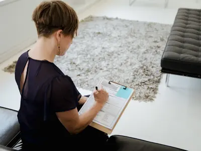 Kobieta w granatowej bluzce wypełnia formularz na podkładce, siedząc na skórzanej kanapie w poczekalni