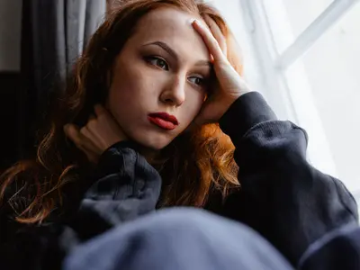Zamyślona rudowłosa kobieta siedzi przy oknie, podpierając głowę dłonią
