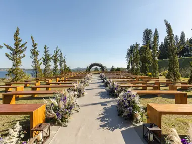 Plenerowa ceremonia ślubna nad jeziorem: drewniane ławki i alejka obsadzona fioletowymi kwiatami