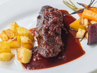 Pieczona wołowina w ciemnym sosie z opiekanymi ziemniakami, marchewką i burakiem na białym talerzu